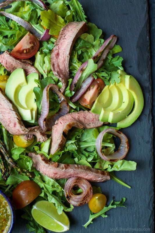 Grilled steak on a bed of arugula salad, topped with tomatoes and avocado on a black slate plate.