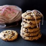 6 chocolate chip cookies tied with pink yarn sitting on a black slate, 1 cookie beside package of cookies.