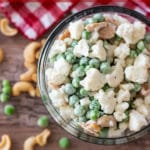 Spoonful of cauliflower and pea salad with cashews in a glass bowl sitting on a white gingham napkin, scattered peas and cashew nuts, silver spoon in bowl.