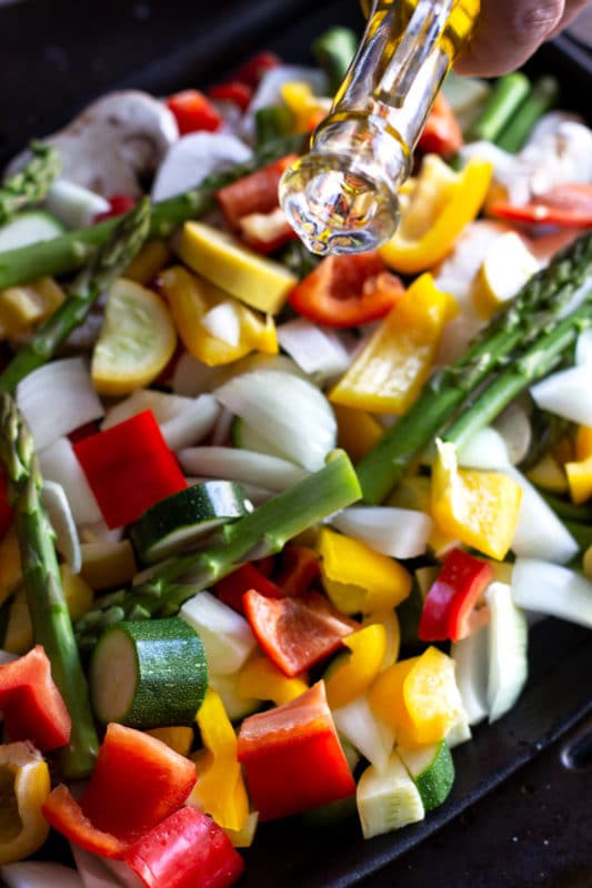 Drizzling olive oil over a pan of chopped vegetables including asparagus, pepper, zucchini and squash.