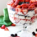 Trifle bowl containing strawberries, blueberries and cheesecake filling sitting on an white table, scattered berries.