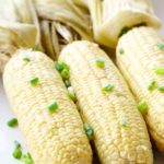 3 cobs of Buttery Smoked Corn on the Cob sitting on a white plate topped with scallions.