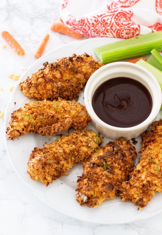 White plate containing 5 air fried gluten free chicken tenders, carrots, celery and BBQ sauce on side.