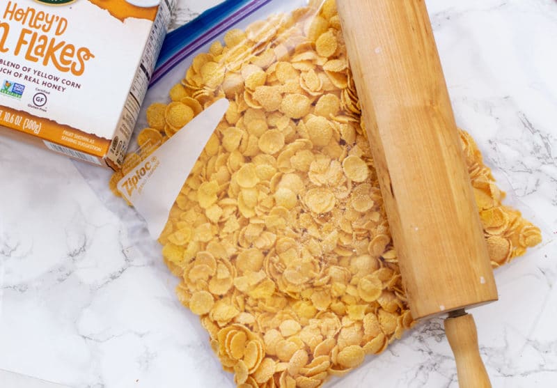 Ziplock bag sitting on a white marble table containing corn flakes, rolling pin and box of corn flakes.