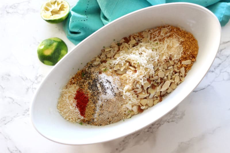 White bowl sitting on a white marbled tabletop containing corn flake mixtures and seasonings, halved limes on table with blue turquoise napkin.