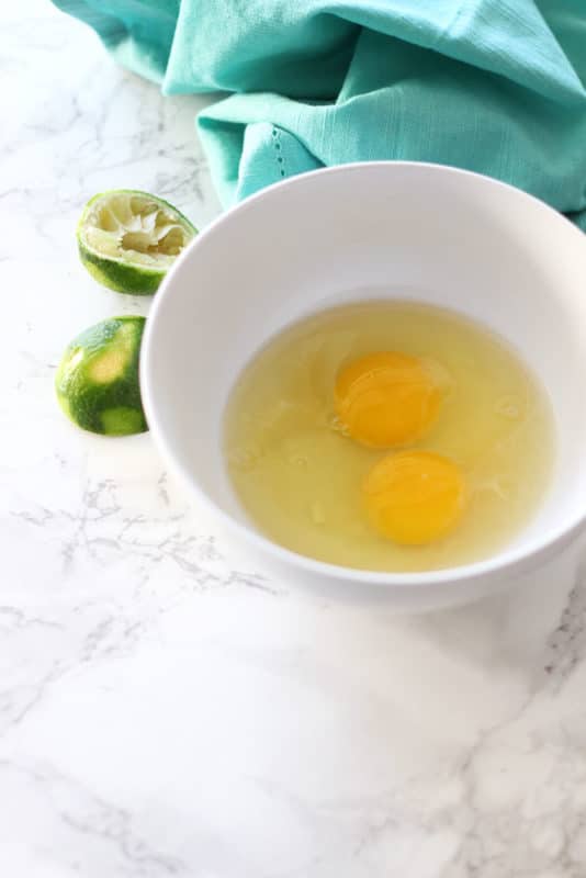 White bowl containing 2 raw eggs sitting on a white marbled table, halved limes and turquoise napkin on side.