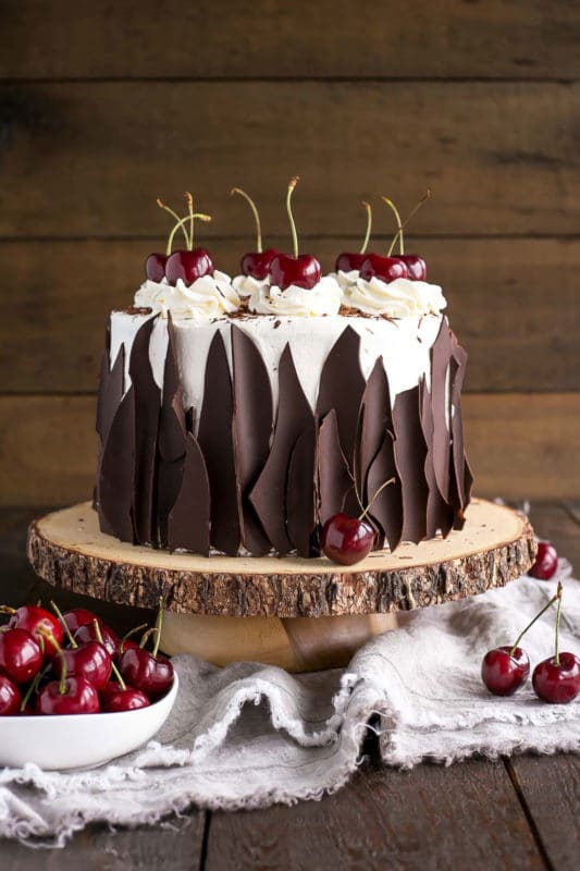 3 tiered Black Forest Cake topped with whipped cream and topped with cherries, bowl of red cherries on side. 