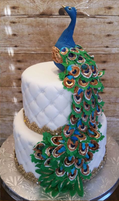 2 tiered white fondant cake topped with a blue peacock, icing feathers cascading down cake. 