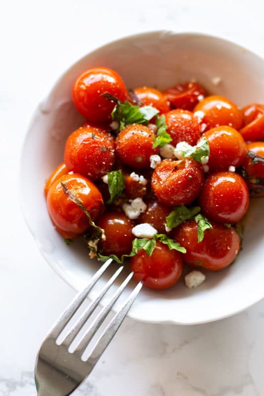 White bowl containing smoked tomatoes topped with goat cheese crumbles and fresh basil, fork in bowl.