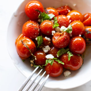 White bowl containing smoked cherry tomatoes topped with goat cheese crumbles and fresh basil, fork in bowl.