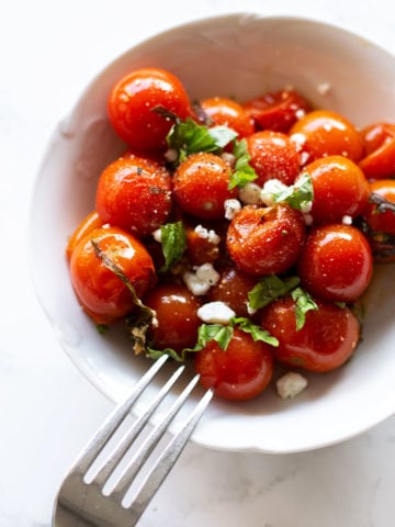 White bowl containing smoked cherry tomatoes topped with goat cheese crumbles and fresh basil, fork in bowl.