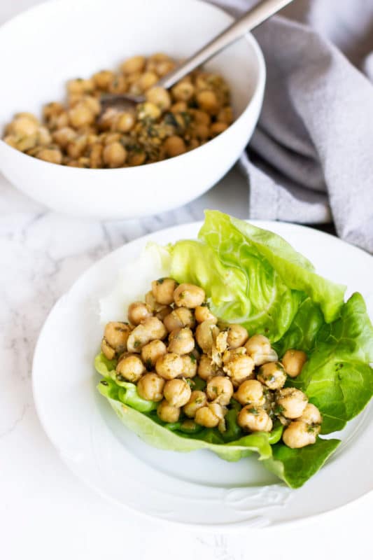 White plate containing a Pesto Chickpea lettuce wrap, bowl of pesto chickpeas on table.