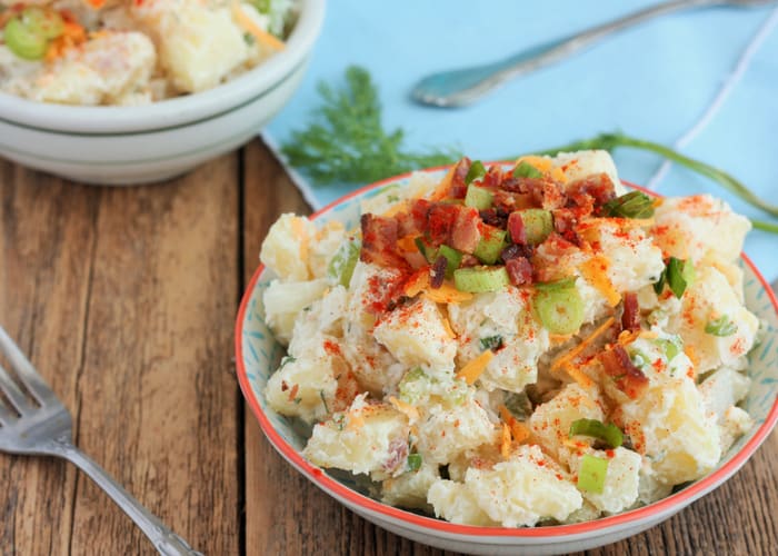 Bowl containing potato salad topped with bacon, cheese and scallions, fork on wooden table.