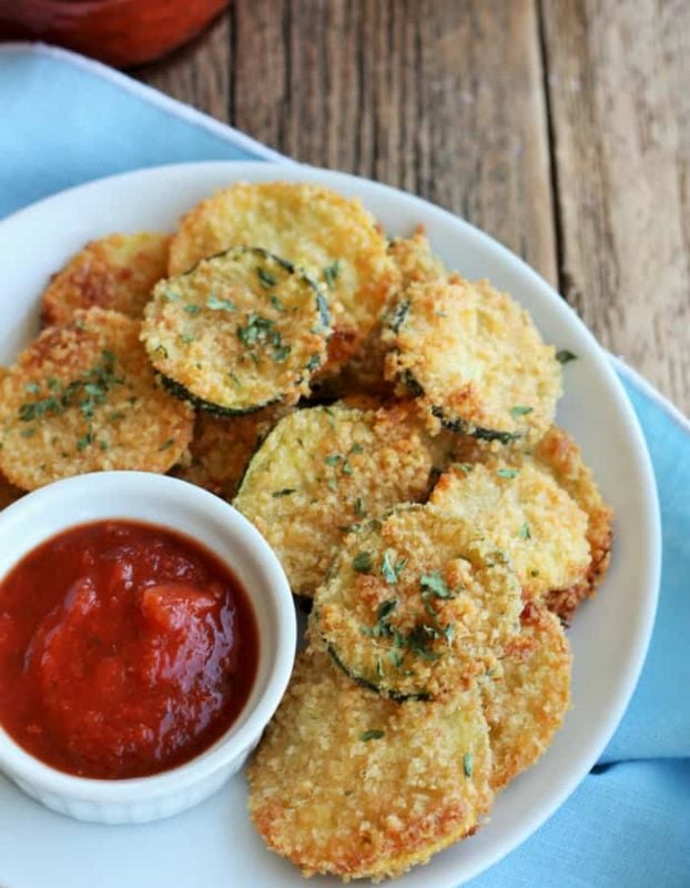 White plate on a wooden table containing 12 Fried Zucchini Chips, side of ketchup.