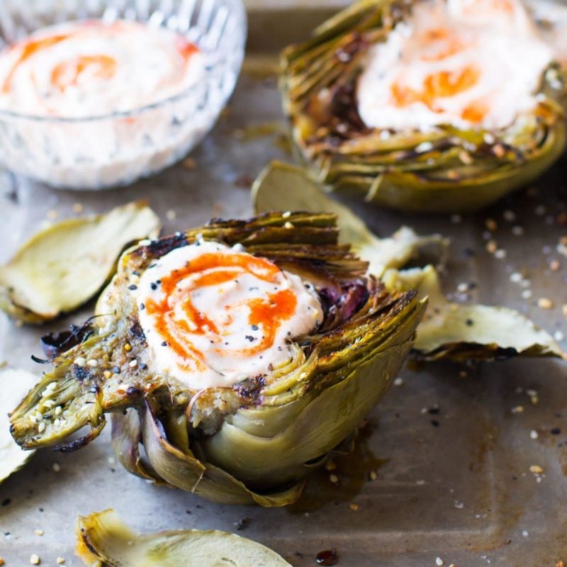 Grilled Artichokes topped with a Sriracha Dipping Sauce on a countertop, dipping sauce on side.