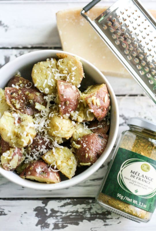 White bowl containing potato salad topped with Parmesan Cheese and Melange De France, grater and Parmesan cheese on white table.