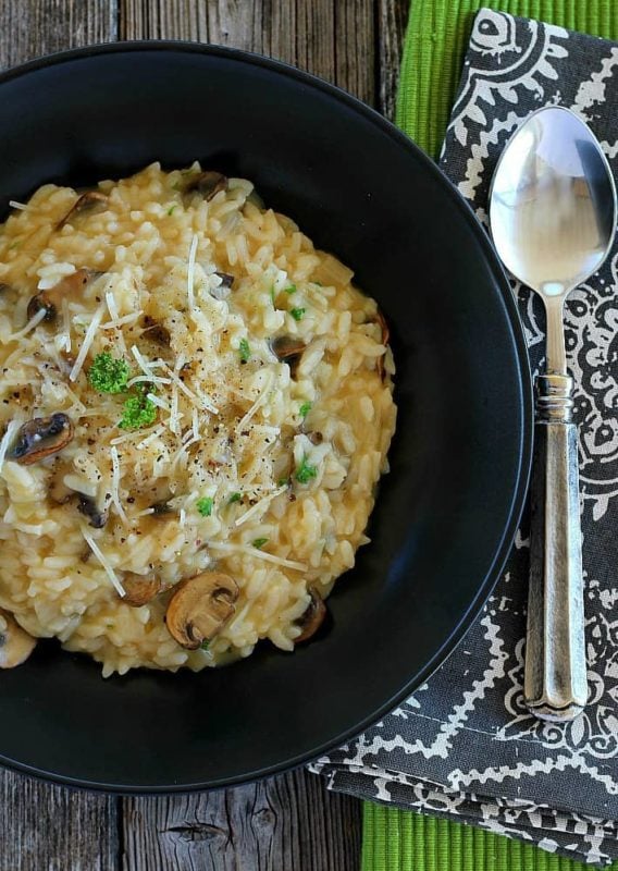 Mushroom risotto in a black bowl topped with mushrooms, scallions and Parmesan cheese, spoon on table.