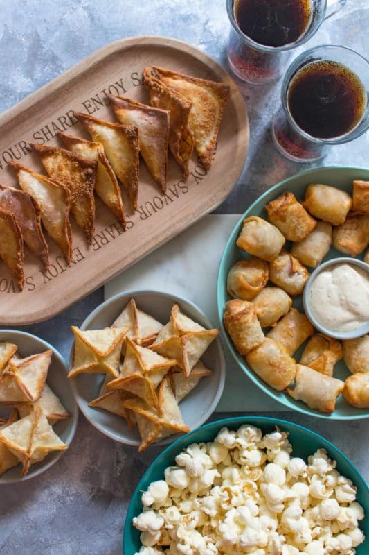 Marble table containing Stuffed Crab Wontons, Sriracha Chicken Wontons and Mini Wonton “Corn Dogs”, bowl of popcorn on side.