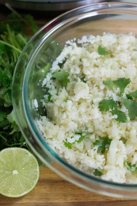 Glass bowl containing cauliflower cilantro rice topped with cilantro, lime on wooden table.