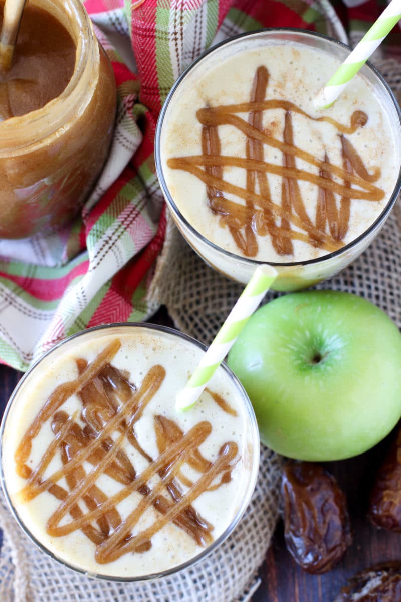 2 Caramel Apple Protein Smoothie in glasses on a table topped with caramel date sauce, green apples and dates on table.