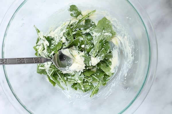 Goat cheese and arugula being stirred by a fork in a glass mixing bowl.