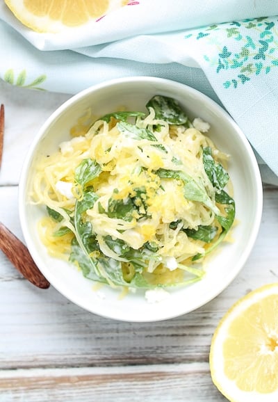 Spaghetti squash with arugula topped with lemon zest in a white bowl with wooden forks and fresh lemons in the background.