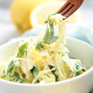 Spaghetti squash with arugula topped with lemon zest in a white bowl with wooden forks and fresh lemons in the background.
