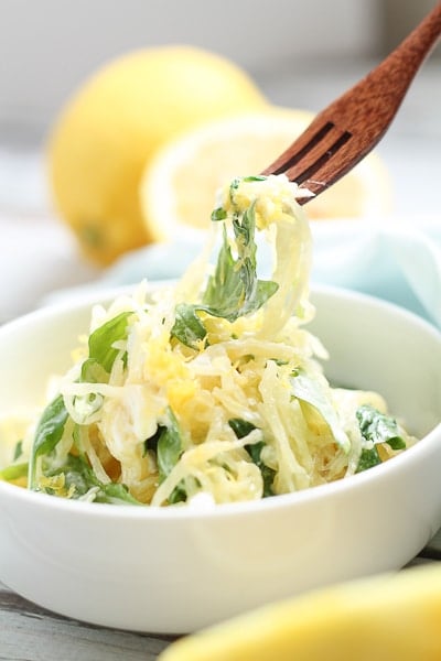 Spaghetti squash with arugula being pulled out of a bowl with a wooden fork with fresh lemons in the background.