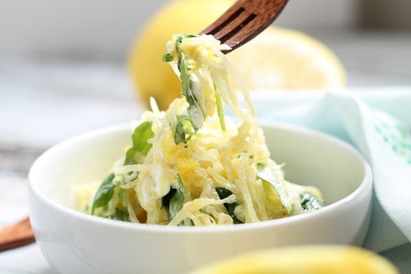 Spaghetti squash with arugula being pulled out of a bowl with a wooden fork with fresh lemons in the background.