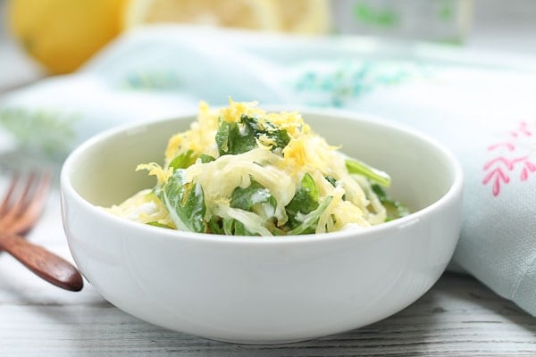 Spaghetti squash with arugula topped with lemon zest in a white bowl with wooden forks and fresh lemons in the background.