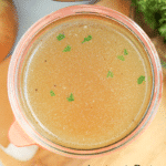 Bone broth in a glass container with parsley on top, garlic cloves and parsley on table.