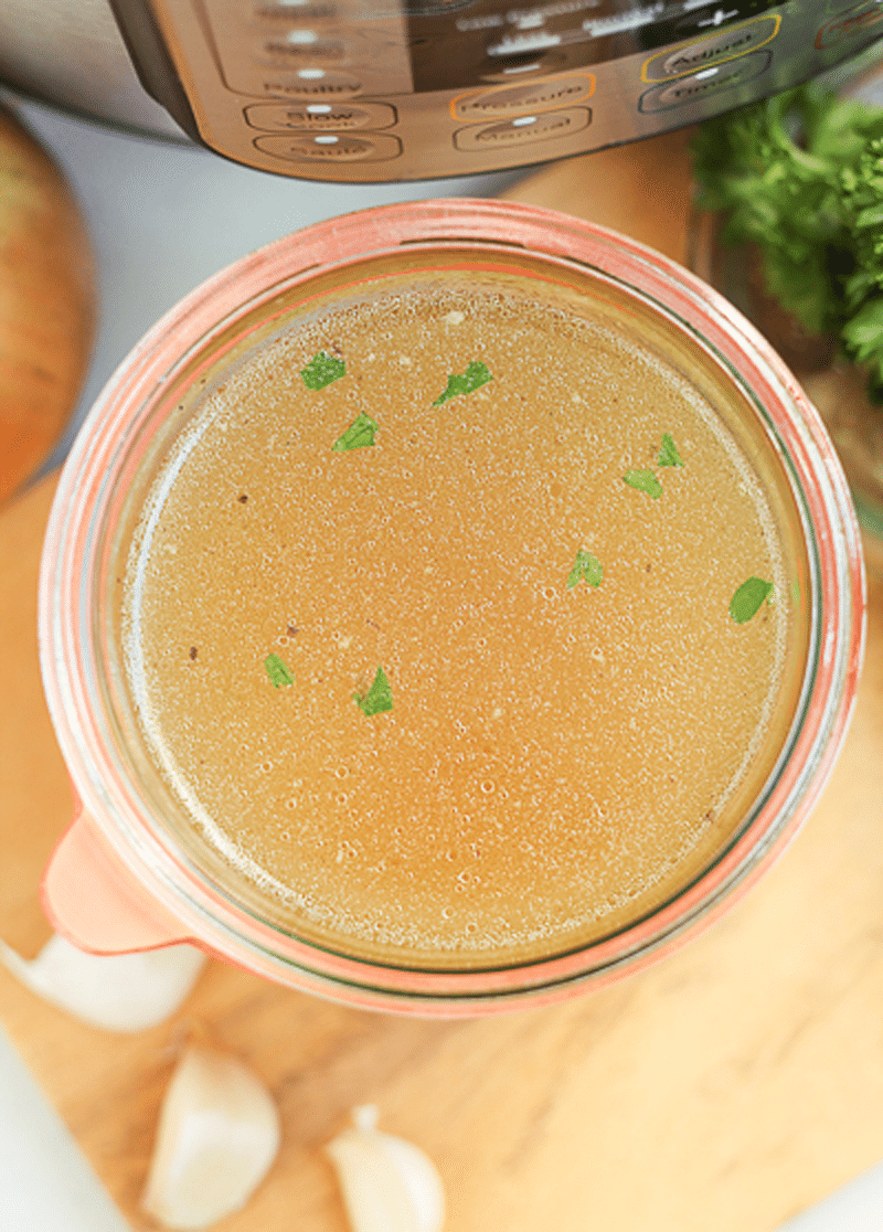 Bone broth in a glass container with parsley on top, garlic cloves and parsley on table. 