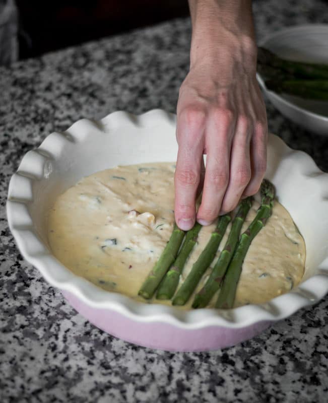 A hand placing asparagus spears neatly on top of a raw quiche mixture.