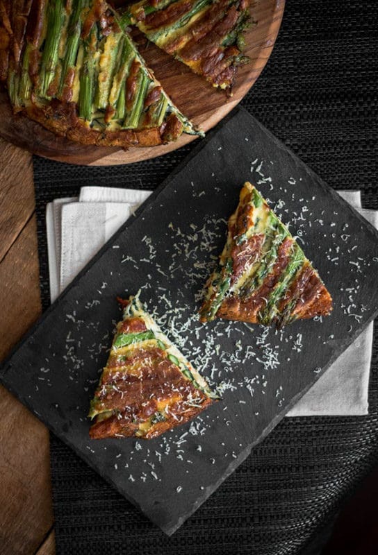 2 slices of an asparagus and tomato filled crustless quiche on a black slate plate topped with Parmesan cheese, quiche on table.