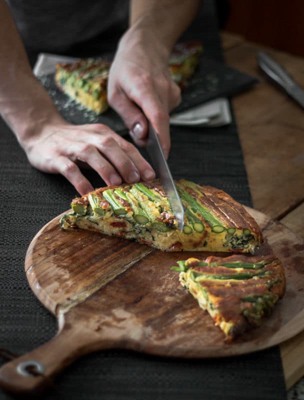 A photo of someone slicing portions out of an asparagus and tomato filled crustless quiche.