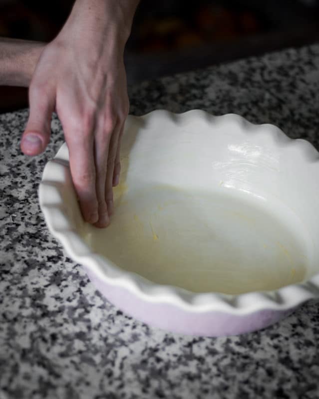 A hand using butter to grease the inside of a pie dish.