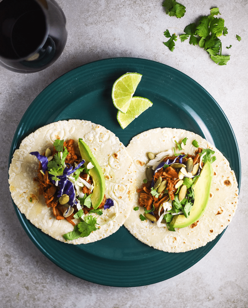 Teal plate containing 2 Jackfruit Tacos paired with a red wine, cilantro on table.