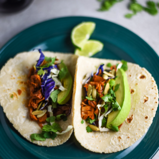 Teal plate on a table containing 2 jackfruit tacos topped with purple cabbage and avocado.