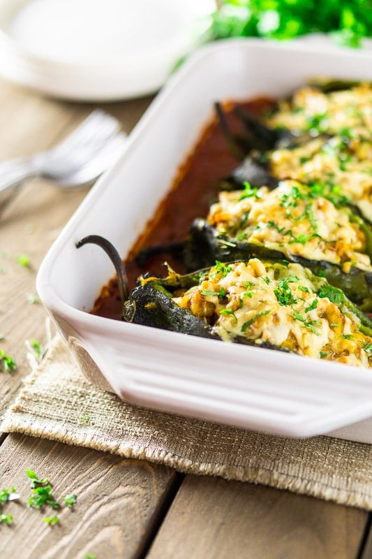 Chorizo-butternut squash chile rellenos in a white pan with ranchero sauce with plates, forks and cilantro in the background.