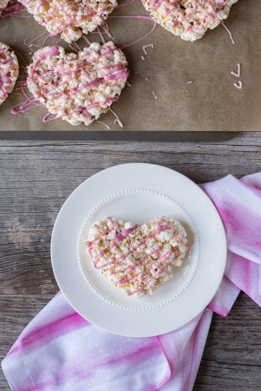 Marshmallow popcorn heart on a white plate with a pink and white towel underneath, hearts on a sheet pan lined with parchment paper toward the top.
