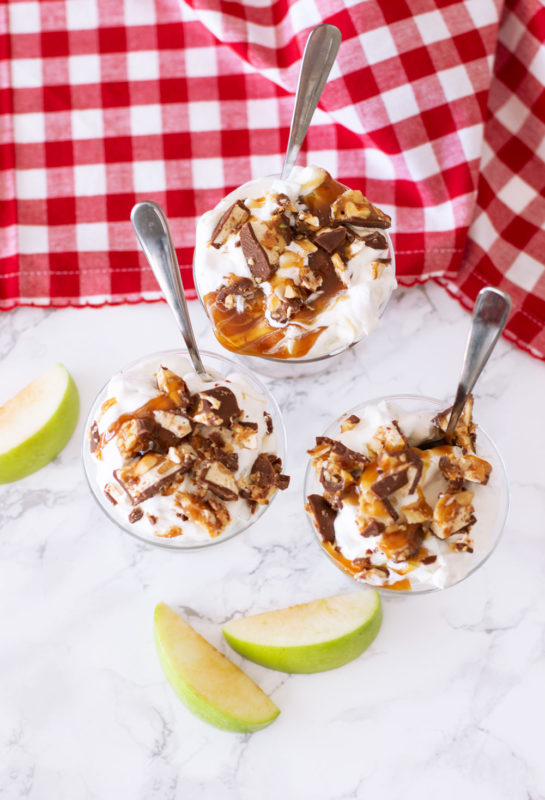 3 parfait glasses containing Apple Snickers Salad, topped with caramel and snickers, apple slices on table.
