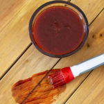 Glass bowl of BBQ sauce sitting on a wooden table, BBQ sauce smeared on table.