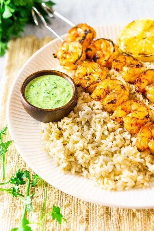 Two shrimp skewers on a bed of rice with a side of summer herb sauce.