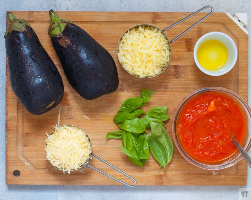 2 eggplants, mozzarella cheese, Parmesan cheese, marinara sauce, olive oil, and fresh basil on a cutting board. 