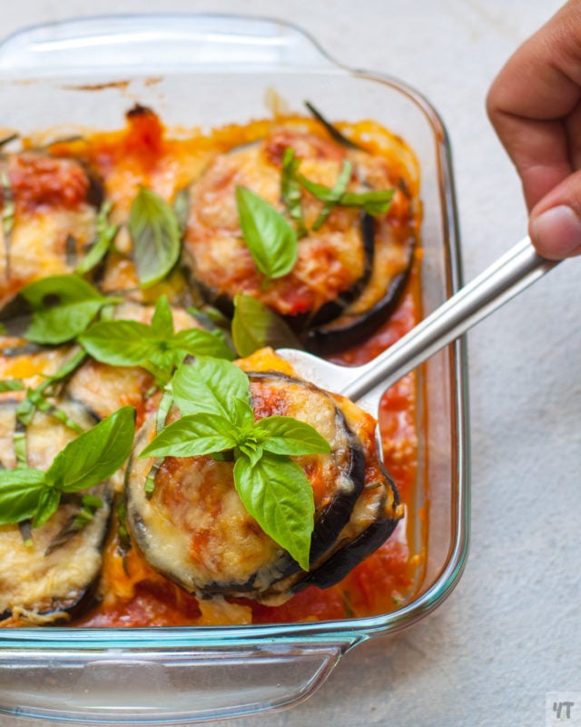 Glass baking dish containing a spoonful of eggplant parmesan topped with cheese and fresh basil.