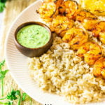 Two shrimp skewers on a bed of rice with a side of summer herb sauce.
