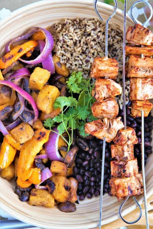 White plate containing grilled jackfruit on skewers, rice, black beans and grilled vegetables.