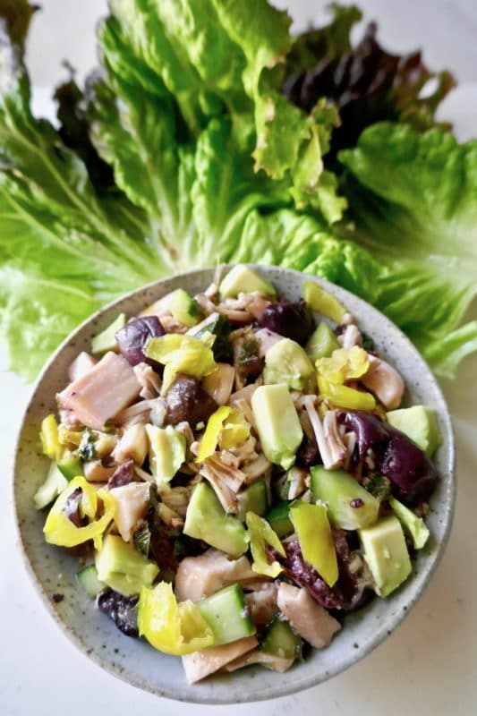 Bowl of salad containing vegan meat, avocado, mixed greens, topped with avocado and kalamata olives.