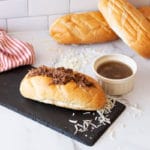 French roll containing shredded beef and cheese, au jus in bowl.