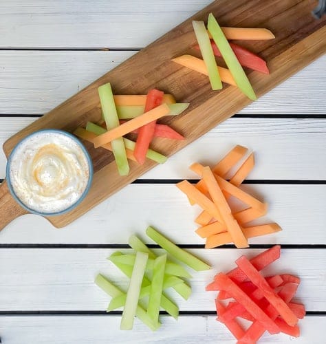 Sticks of melon in piles on cutting board with yogurt dip on the side.